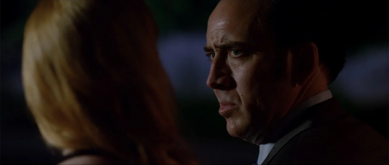 Rage (2014) movie still: closeup — An outdoor night scene shows a man in a dark suit standing outside. The man stares intently at a person whose…