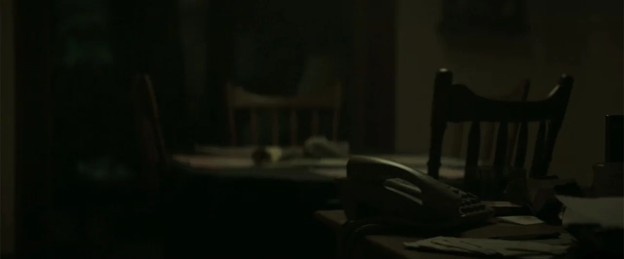 Crown Heights (2017) movie still: medium — A dimly lit room contains a wooden table with chairs and a landline telephone. The furniture is barely…