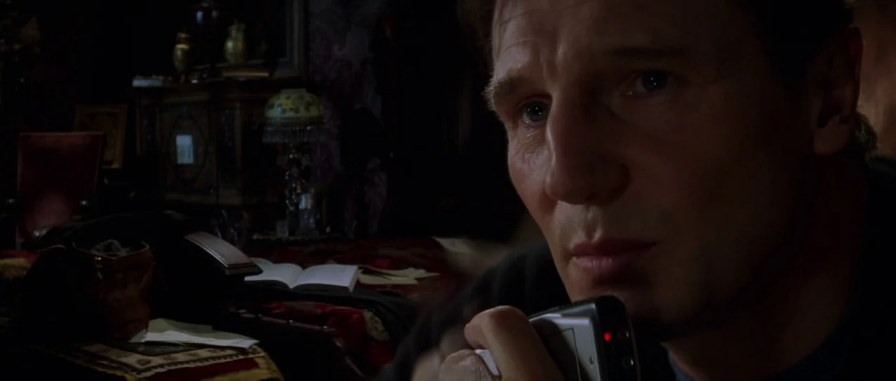 The Haunting (1999) movie still: closeup — A dark bedroom features a vintage lamp and an open book on a bed. The man holds a small digital device…