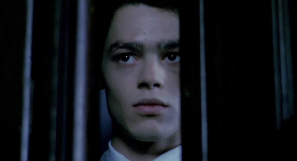 In a Glass Cage (1986) movie still: closeup — A man stands behind heavy iron bars in a dark room. The subject looks forward with a neutral…