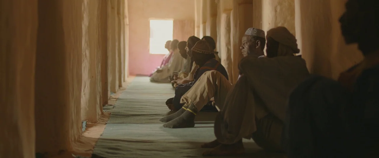 Timbuktu (2014) movie still: medium — A long, narrow room with thick, rough-textured mud walls and mat-covered flooring. Several men sit quietly in…