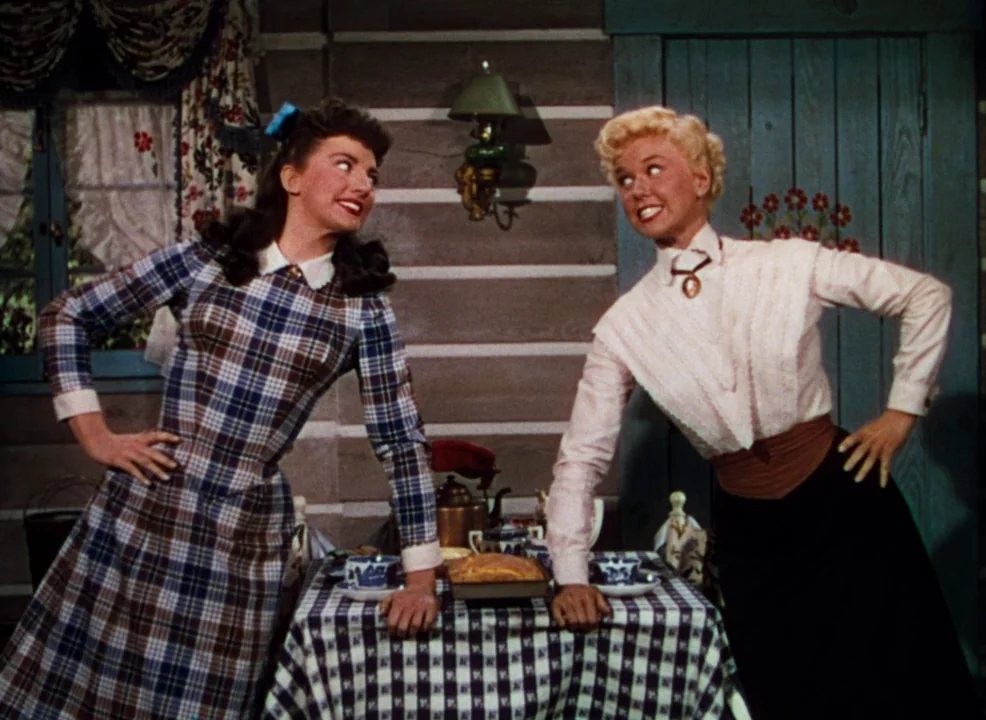 Calamity Jane (1953) movie still: medium — A rustic log-walled interior features a table set for tea with a plaid cloth. Two women in vintage…