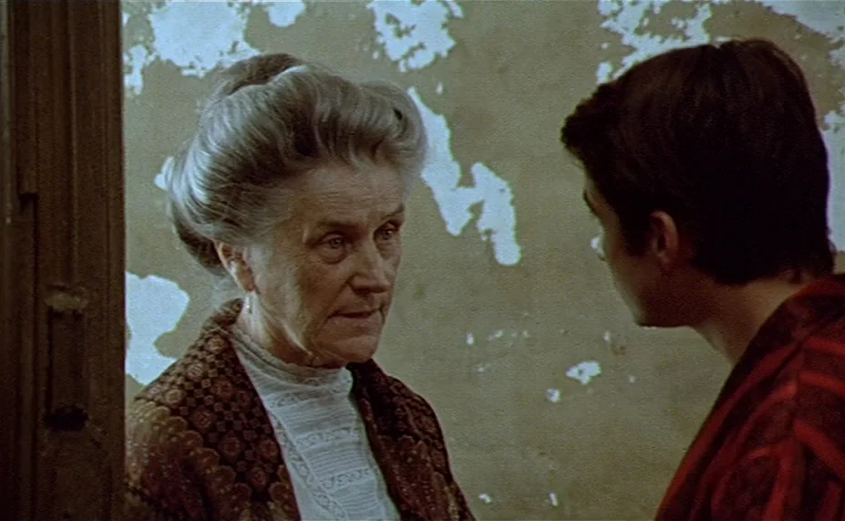 Two English Girls (1971) movie still: medium — A dimly lit indoor space features a wall with peeling light-colored paint. An elderly woman faces a…