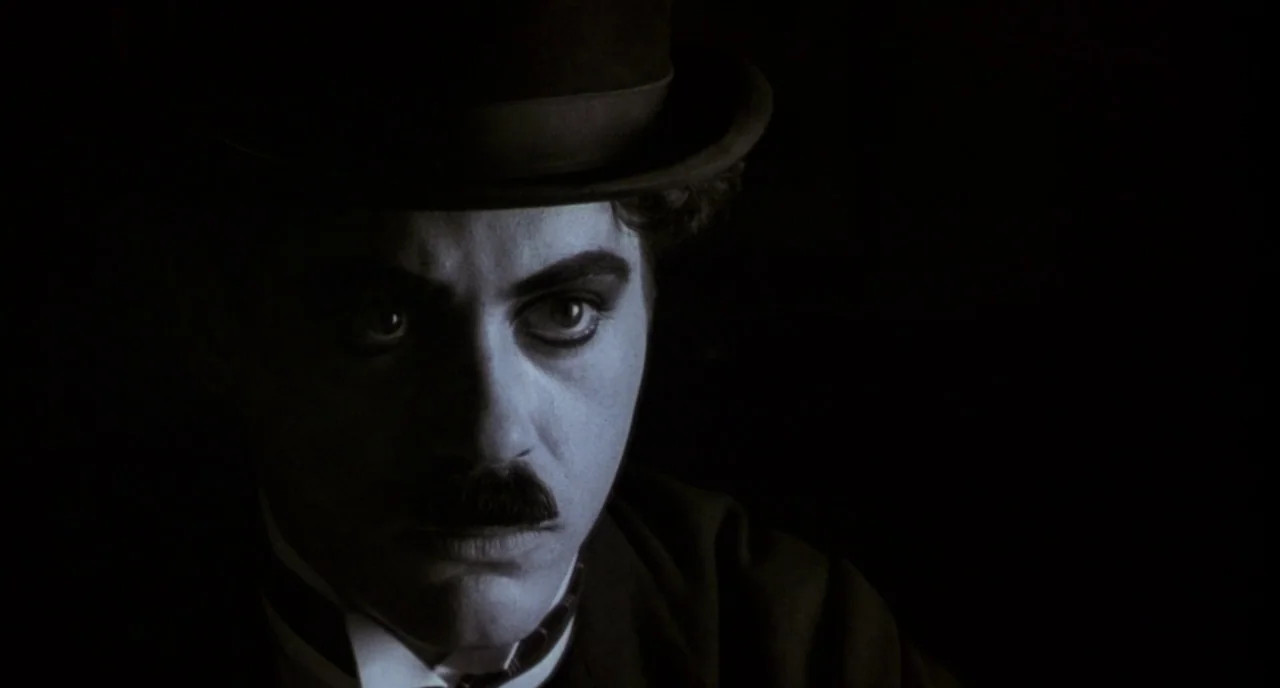 Chaplin (1992) movie still: closeup — A man wearing a bowler hat stands against an entirely black void. He stares forward with heavy stage makeup…