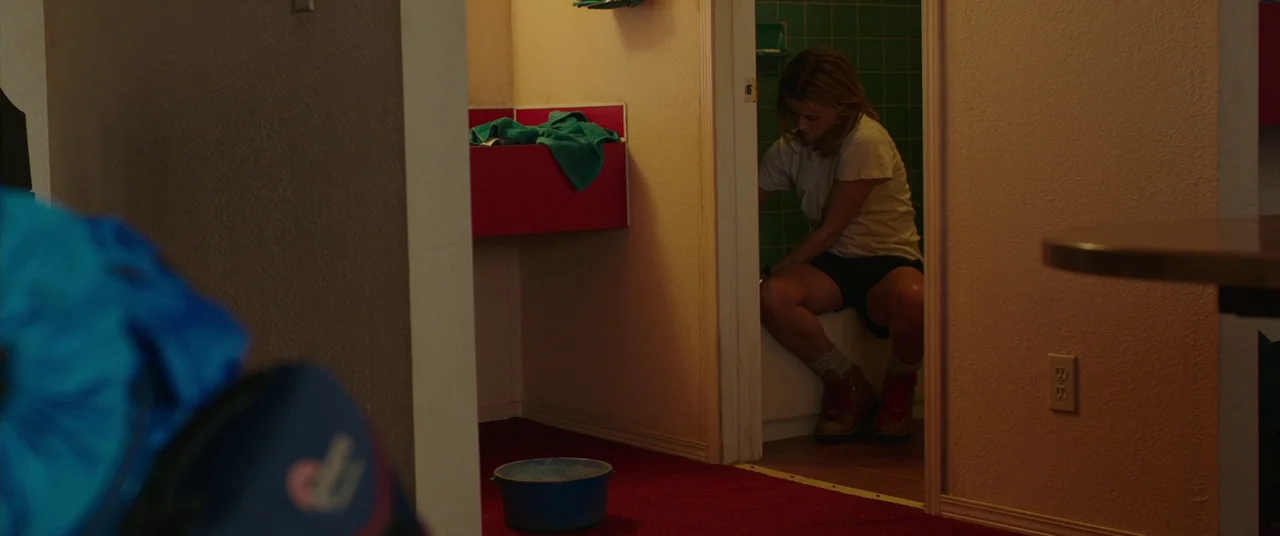 Wild (2014) movie still: wide — A small motel bathroom features dark green tiled walls and a red carpeted area. A woman in a t-shirt and hiking…