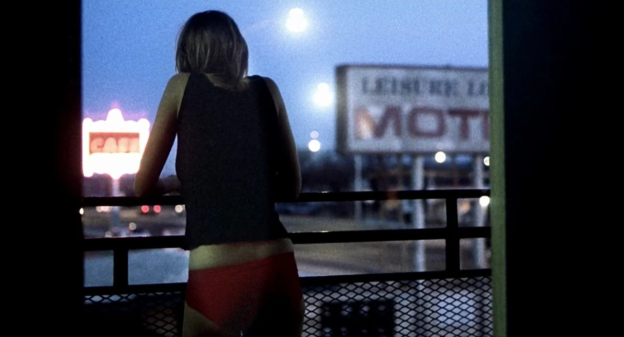 21 Grams (2003) movie still: medium — A woman stands on a dark metal motel balcony at night. In the background, illuminated neon signs for a cafe…