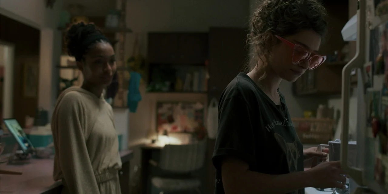 Sitting in Bars with Cake (2023) movie still: medium — A dimly lit indoor space with blurred furniture and home items in the background. Two young…