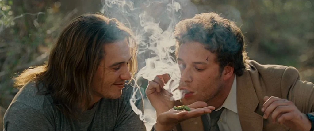 Pineapple Express (2008) movie still: medium — Two men sit in a brushy field with out-of-focus natural foliage in the background. One man blows…