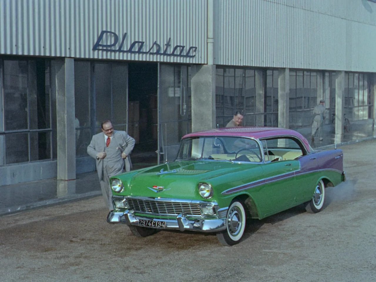 Mon Oncle (1958) movie still: wide — A modern industrial building with a large Plastac sign sits behind a parked car. A man in a grey suit steps out…
