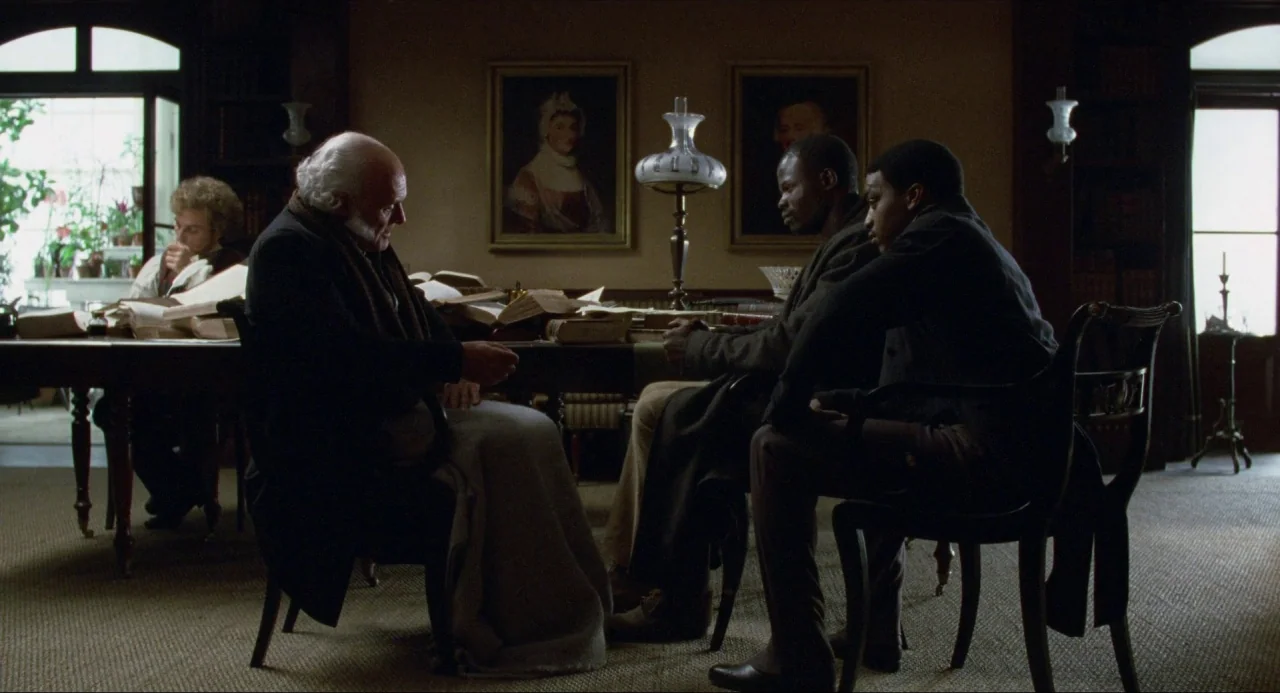Amistad (1997) movie still: wide — An 1800s wood-paneled room with books, framed portraits, and a large central table. Three men are seated in…