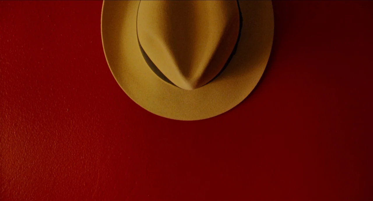 Dick Tracy (1990) movie still: closeup — A tan felt fedora hangs centered on a solid matte-red wall. The texture of the hat material contrasts with…