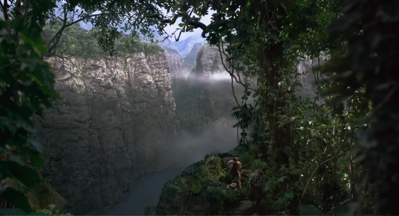 Jurassic Park III (2001) movie still: wide — A deep canyon features steep rock walls and dense vegetation overlooking a river. Two figures stand on…