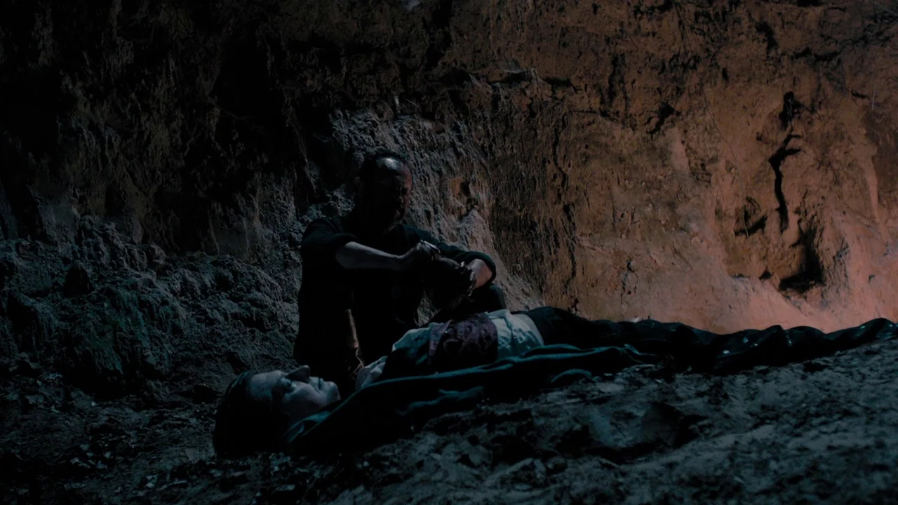 The Field Guide to Evil (2018) movie still: medium — A rugged cave interior with rough, earthy rock walls forms a dark, tight space. A seated man…