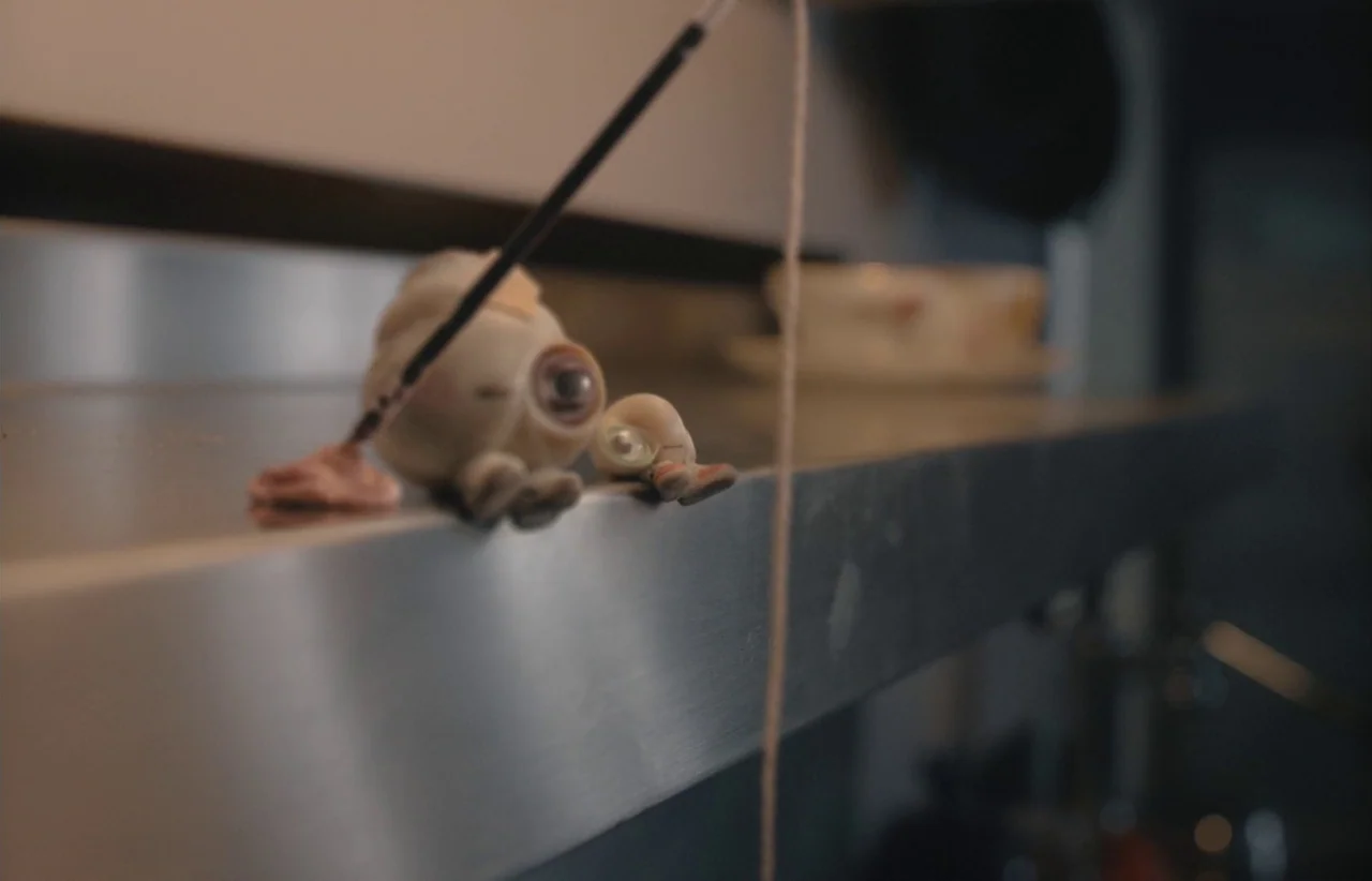 Marcel the Shell with Shoes On (2021) movie still: closeup — A polished metal edge of a shelf supports two small, pale eyeball-shaped figurines. The…