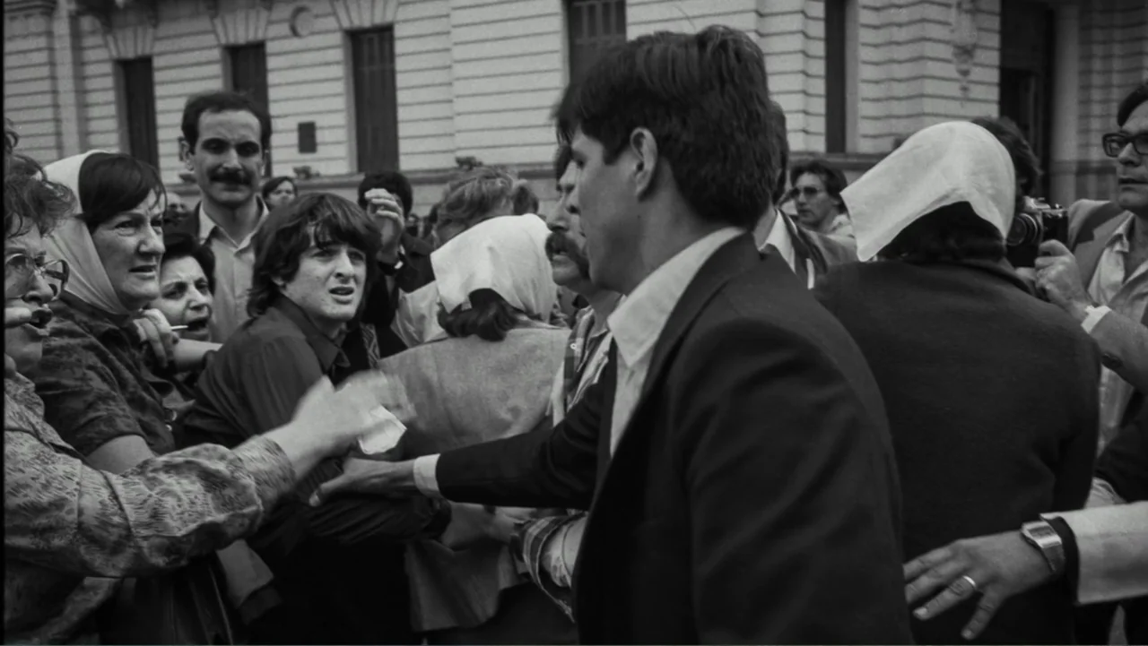 Transfers (2024) movie still: medium — A group of people gathers in front of a classical stone building with windows. A man in a suit pushes through…