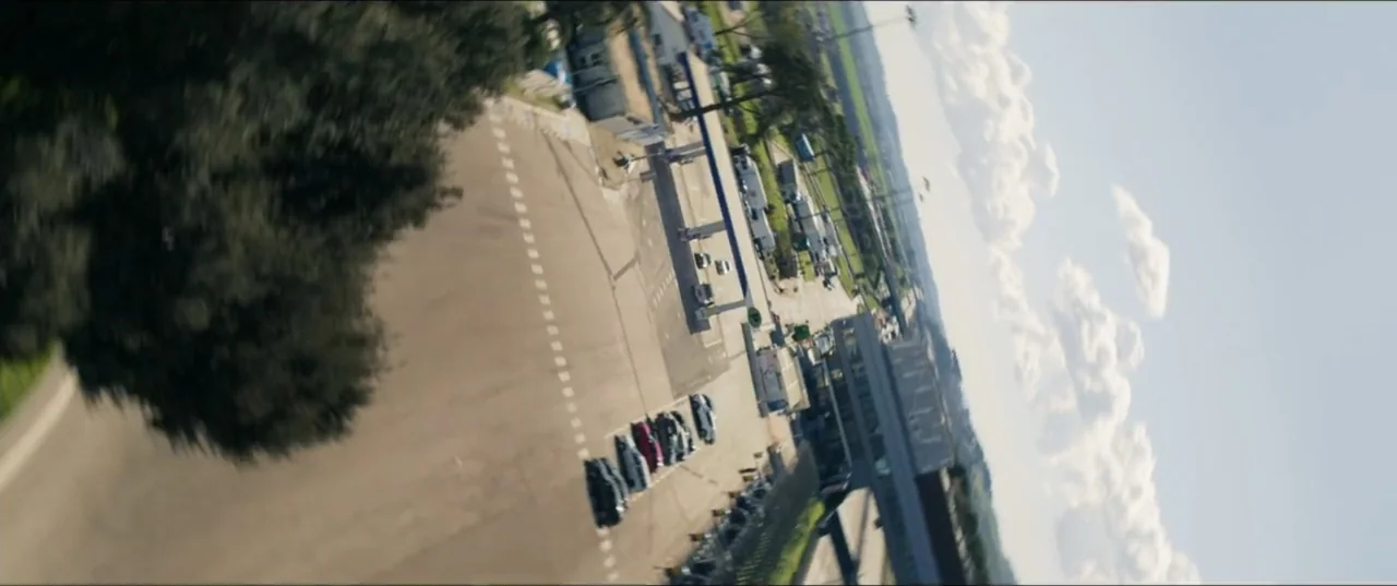 Smurfs (2025) movie still: establishing — A high-angle view looks down at a gas station parking lot from above. Several cars are parked in…