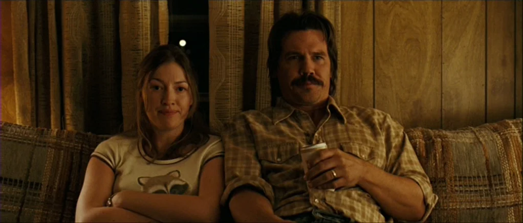 No Country For Old Men movie still: medium — A domestic space with wood-paneled walls and textured curtains behind a plaid sofa. A man and a woman…