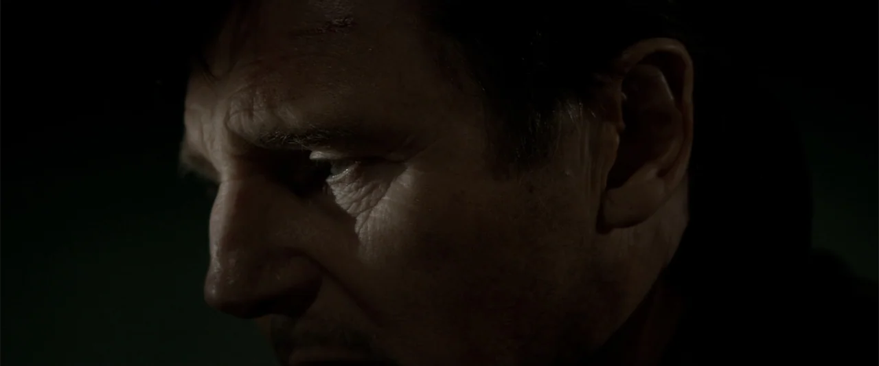 A Walk Among the Tombstones (2014) movie still: closeup — A dark, dimly lit interior space surrounds a man in close proximity. A man with weathered…