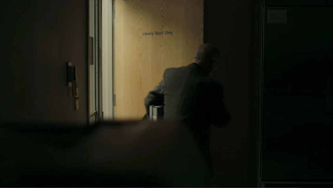 Alabama Snake (2020) movie still: medium — A dimly lit indoor corridor features a light wooden door labeled Library Staff Only. A man in a dark…