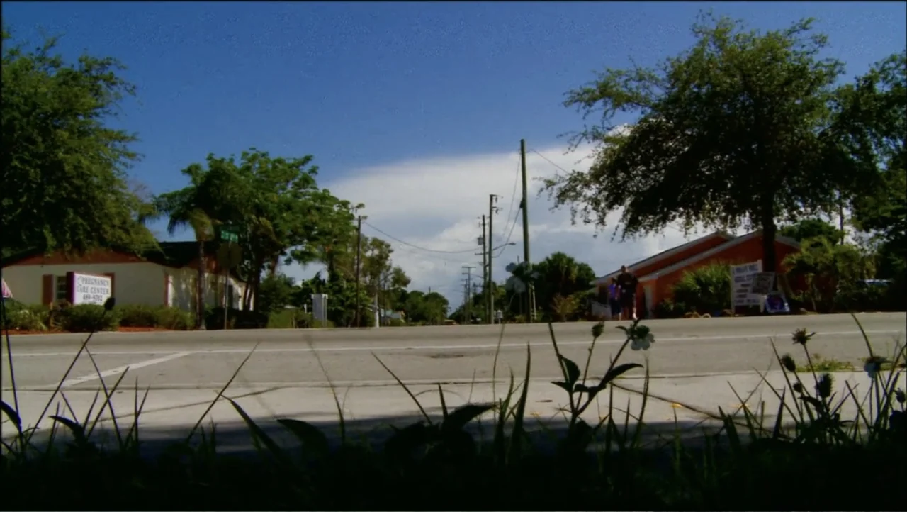 12th & Delaware (2010) movie still: establishing — A quiet suburban road in daylight with trees and single-story buildings on both sides. The camera…