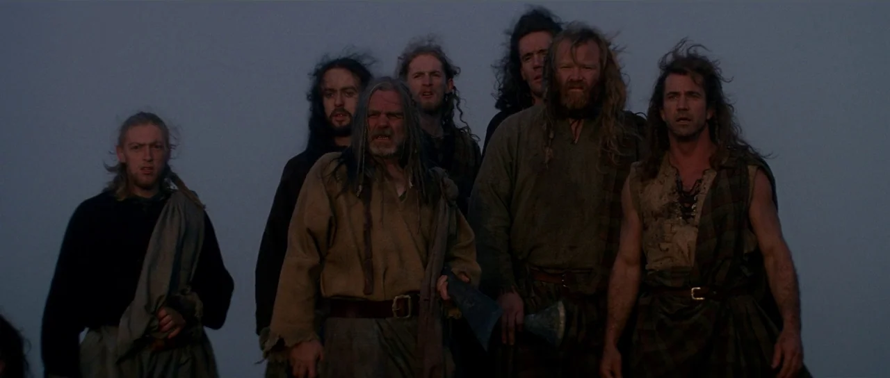 Braveheart (1995) movie still: medium — A group of men stand together against a featureless grey, overcast sky. The men look forward, appearing…