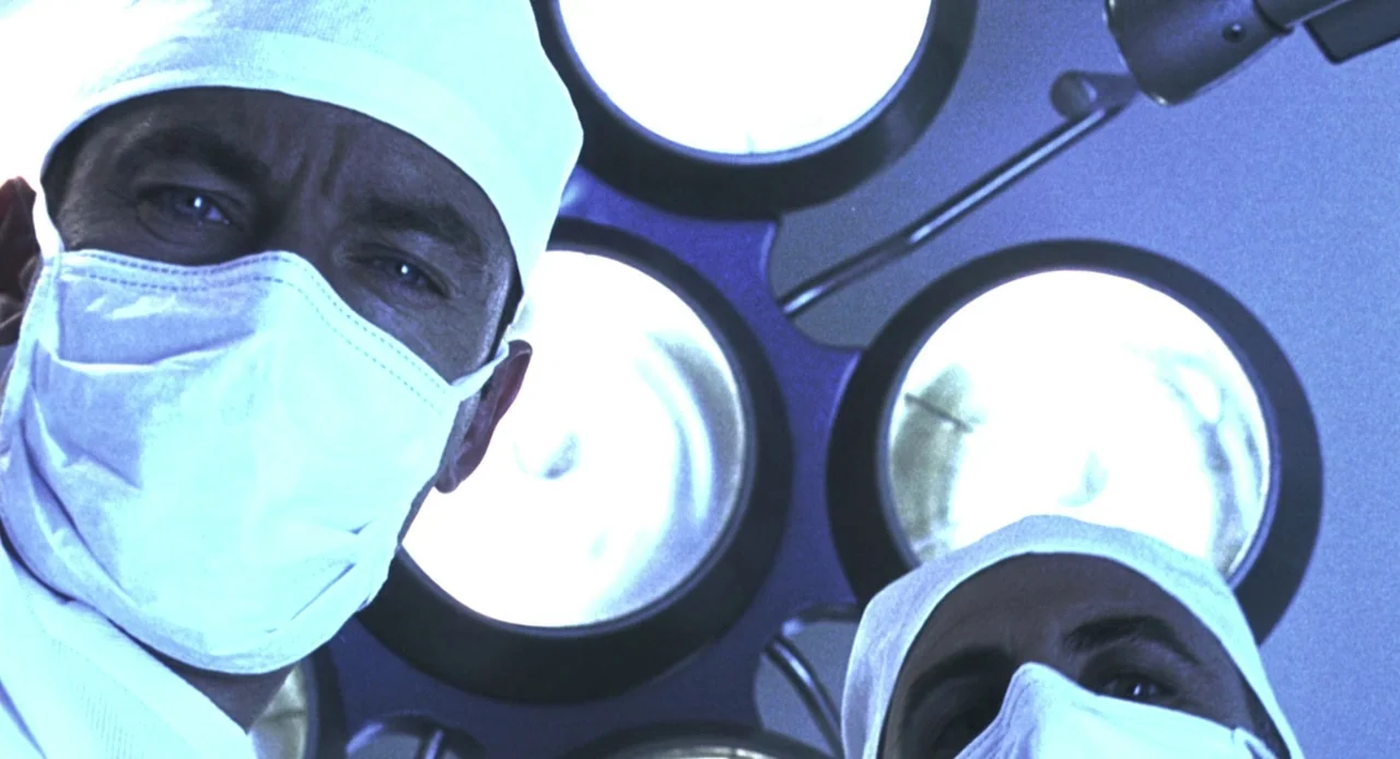 Resident Evil (2002) movie still: closeup — A clinical surgical environment featuring powerful multi-lamp overhead lights. Two medical professionals…