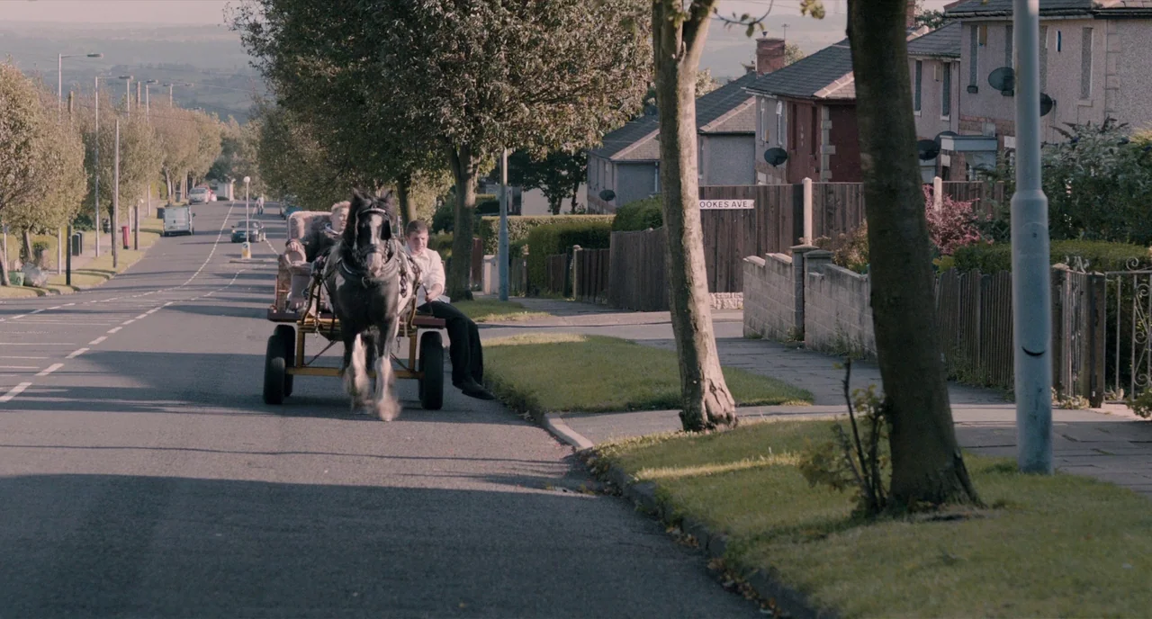 The Selfish Giant (2013) movie still: wide — A residential road in a suburban area features a paved street and paved sidewalks. Two young men ride…
