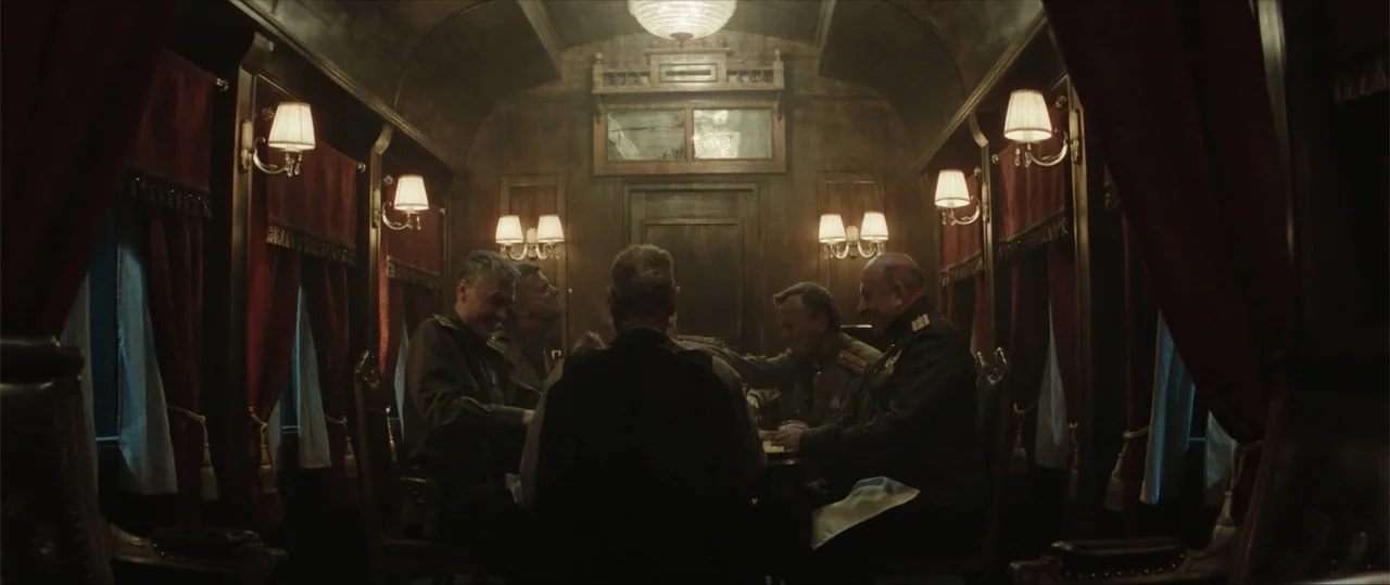 Sisu: Road to Revenge (2025) movie still: medium — An interior train carriage features dark wood paneling and red fabric window drapes. Several men…