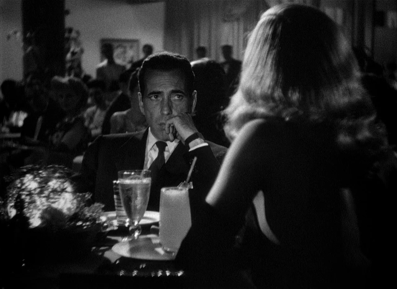 Dead Reckoning (1946) movie still: medium — A dark, crowded room features blurred figures in the background beyond the table. A man in a suit looks…