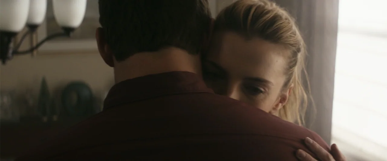 The Tomorrow War (2021) movie still: closeup — A dimly lit interior space features two figures in close proximity. A man seen from the back embraces…