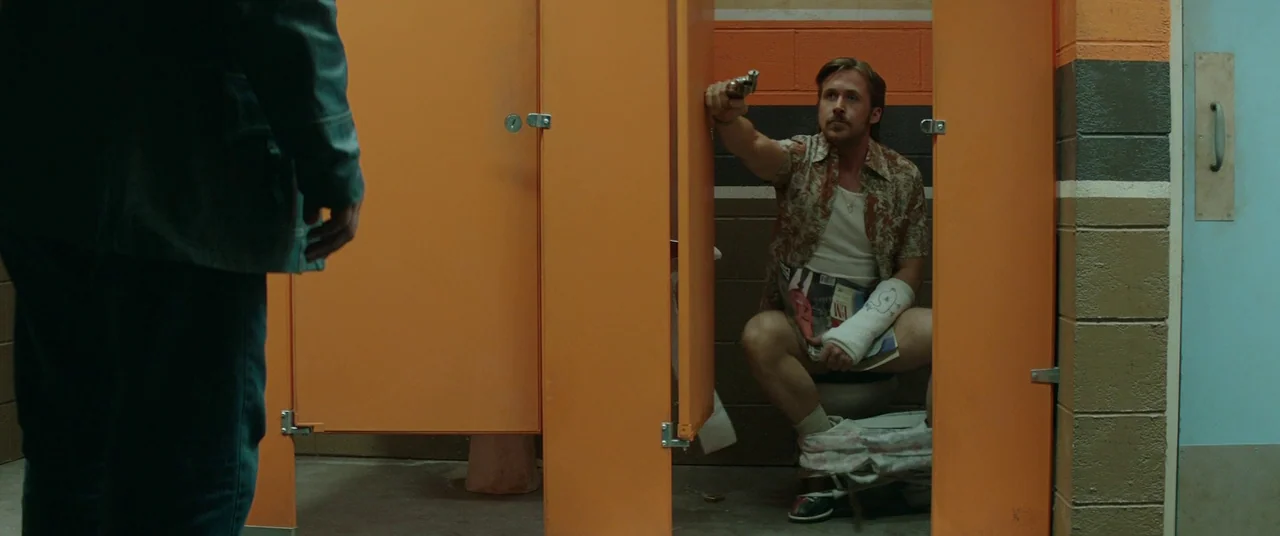 The Nice Guys (2016) movie still: wide — A public restroom features bright orange stall doors and multi-toned block walls. A man with a broken wrist…