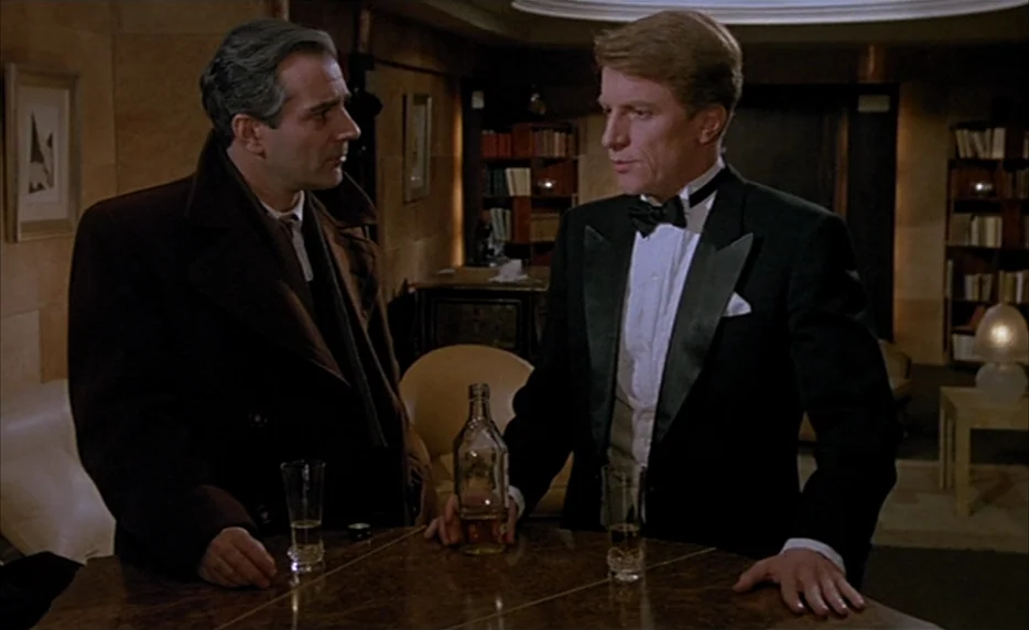 Mélo (1986) movie still: medium — A dimly lit room features a wooden table with drinks in a lounge area. Two men in formal attire stand together…
