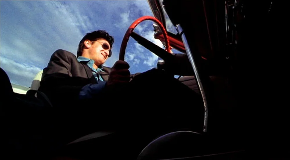 U-Turn (1997) movie still: medium — The interior of a vehicle with red steering wheel detailing under a blue sky. A man sits in the driver seat…