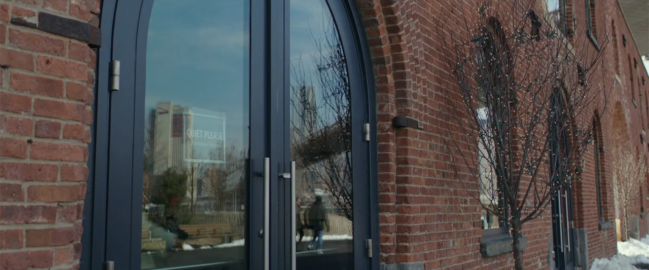 Collateral Beauty (2016) movie still: medium — An arched double-glass door is set into a rustic red brick building wall. A sign on the glass reads…