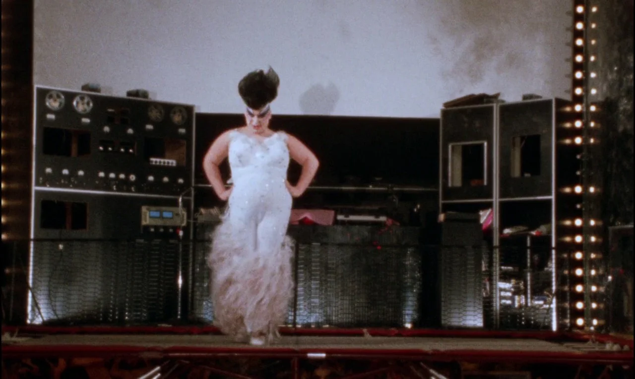 Female Trouble (1974) movie still: fullbody — A performer stands center stage in front of large vintage reel-to-reel tape machine equipment. The…