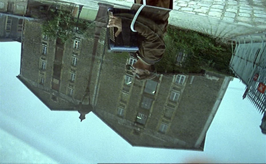 Diva (1981) movie still: medium — The image shows a reflection of a man playing an accordion in a window. The background shows a multi-story stone…