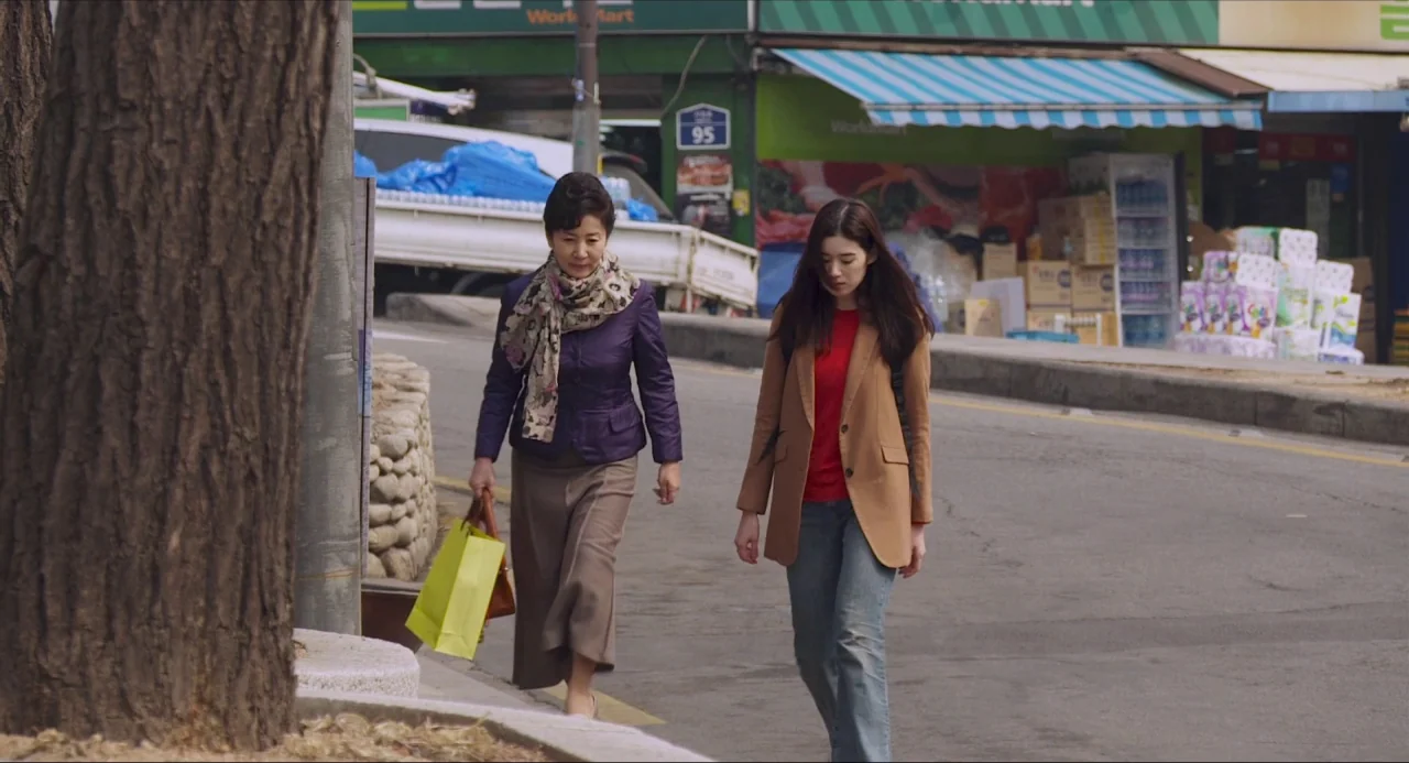 Nobody’s Daughter Haewon (2013) movie still: fullbody — Two women walk along the paved edge of a road beside a tree trunk. Shops with green signage…