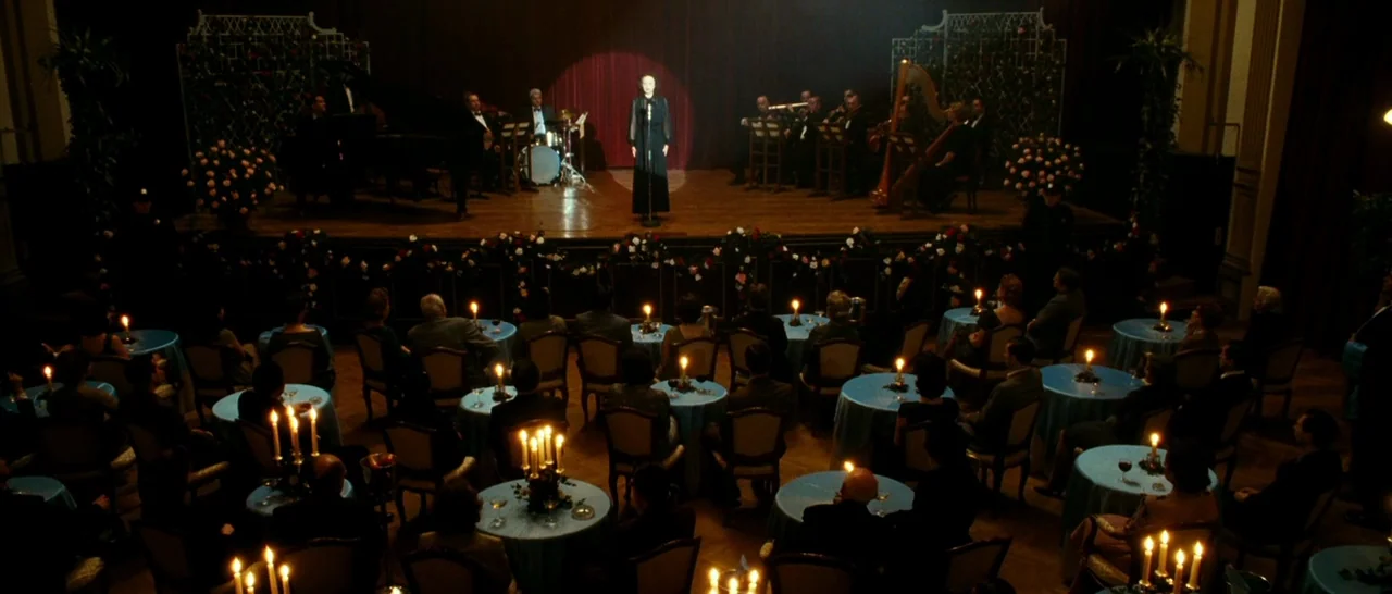La Vie En Rose (2007) movie still: wide — A dark ballroom is filled with candlelit tables and a band on a wooden stage. A woman stands in a…