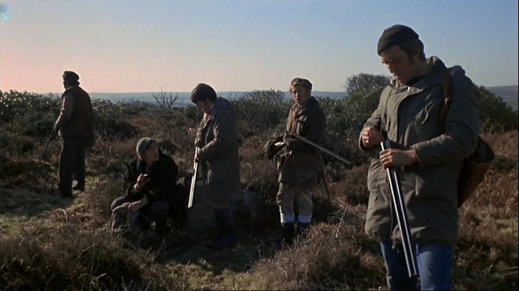 Straw Dogs (1971) movie still: fullbody — A group of men in outdoor hunting gear stand across a barren, brush-covered heath. The men handle…