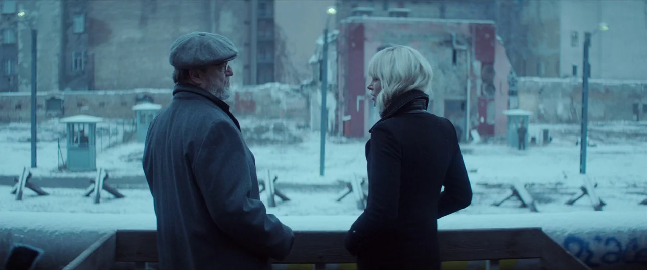 Atomic Blonde (2017) movie still: medium — A snowy, desolate border crossing with anti-tank obstacles and guard booths in the distance. An older man…