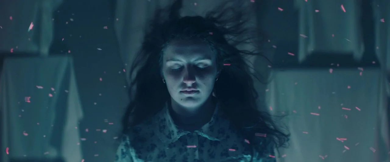 Extra Ordinary (2019) movie still: closeup — A woman stands in a dimly lit, dark space with hanging fabric elements in back. Red glowing particles…