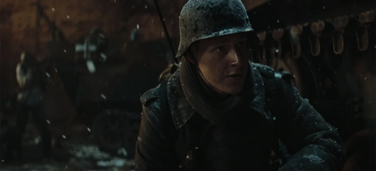 The Tank (2025) movie still: medium — A snowy battlefield with a soldier positioned near a large heavy military vehicle. The soldier wears a combat…
