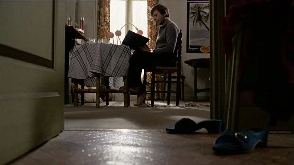 The Tenant (1976) movie still: medium — A furnished room with floral drapes and a poster of a palm tree. A man sits at a small table reading a…