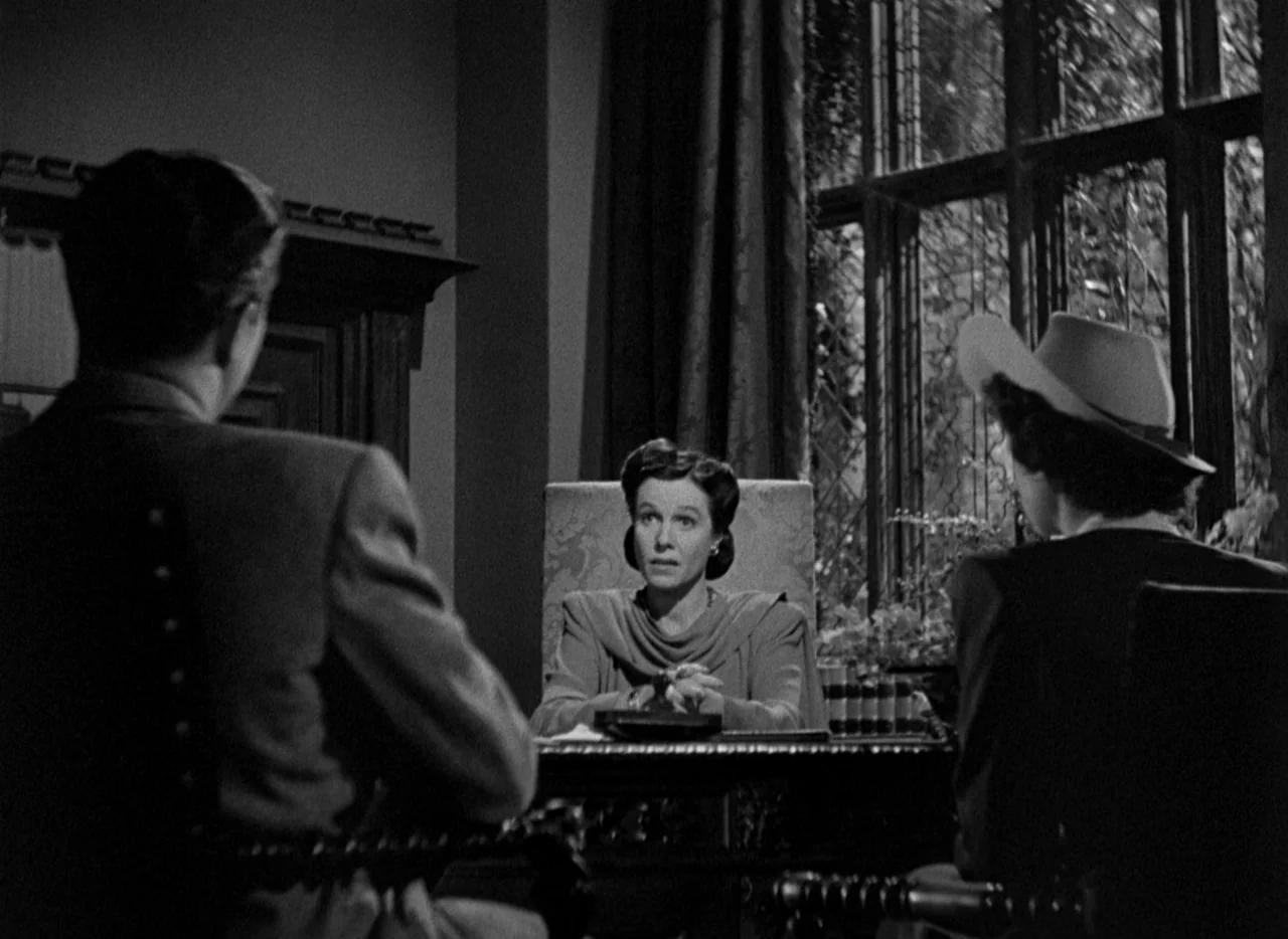 The Uninvited (1944) movie still: medium — A dark wood-paneled office features a large leaded glass window behind a central desk. A woman sits at…