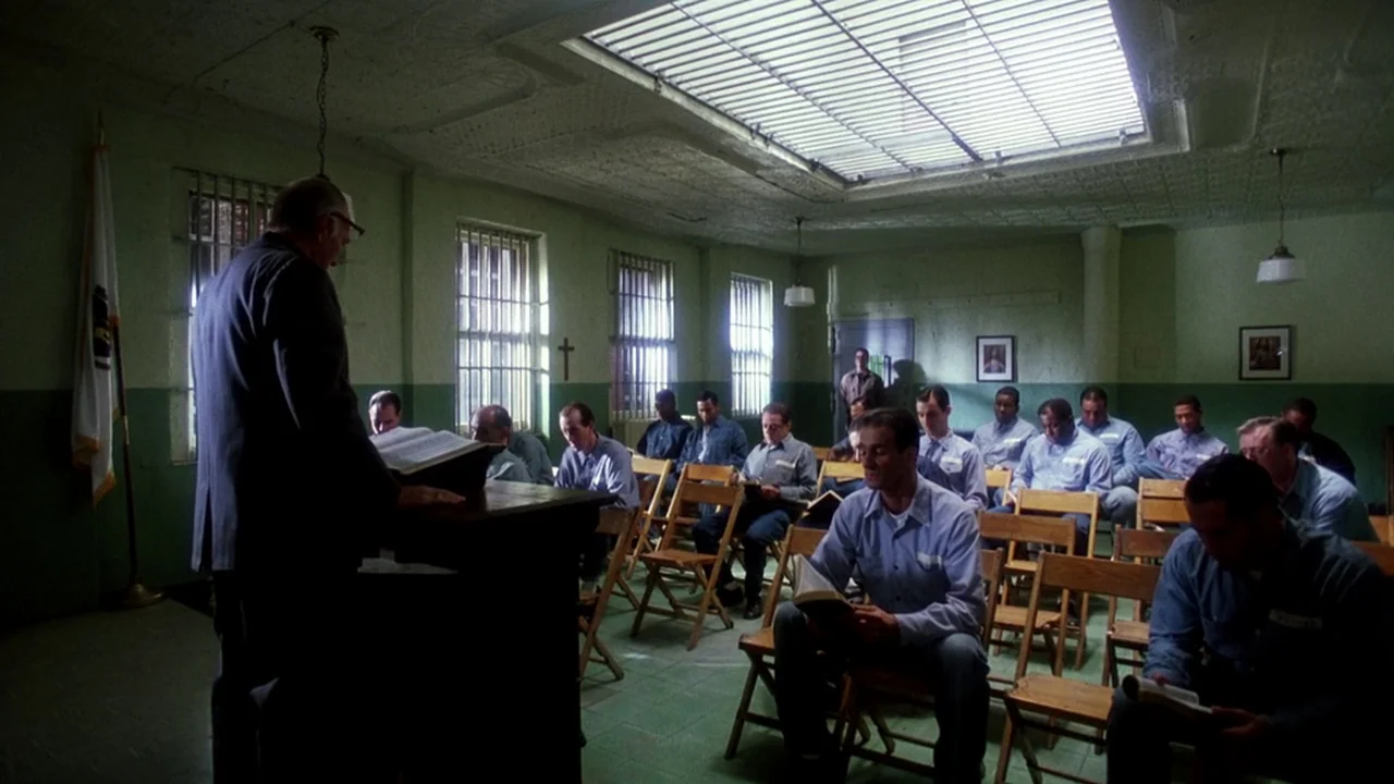 Malcolm X (1992) movie still: wide — A prison chapel features green walls, rows of wooden chairs, and barred windows. A group of men in light blue…