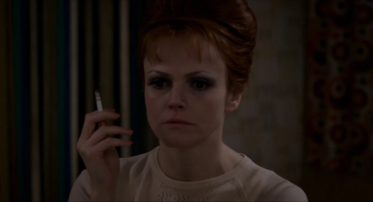 The Falling movie still: closeup — A woman with a high hairstyle sits in a dimly lit interior space. She holds a lit cigarette between her fingers…