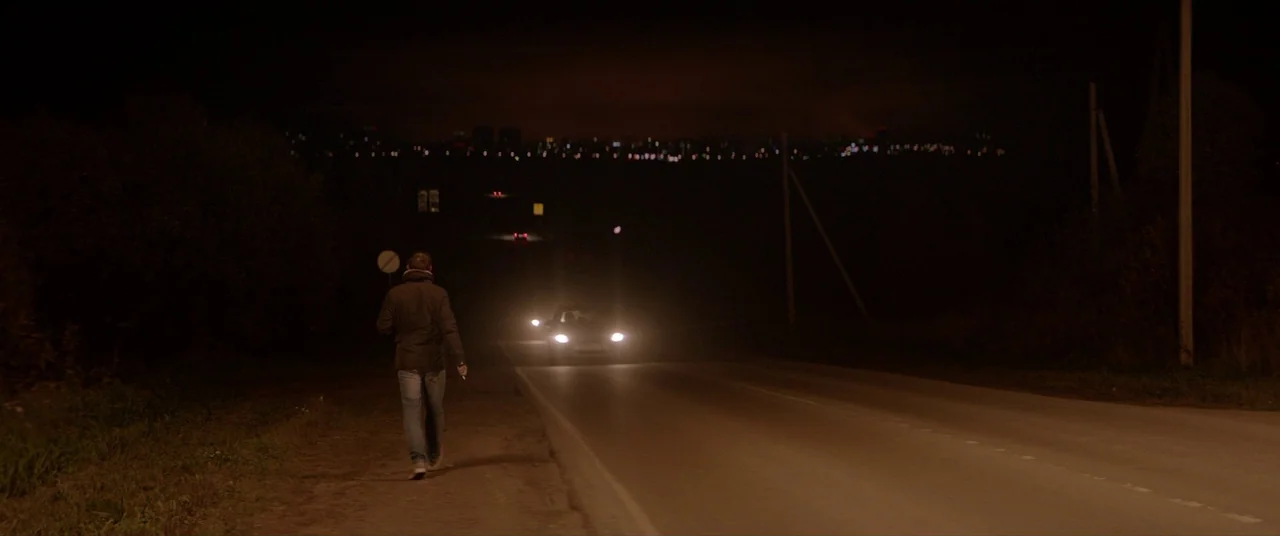Loveless (2017) movie still: wide — A dark, empty paved road is lined with trees under a night sky. A man walks on the shoulder while a car…