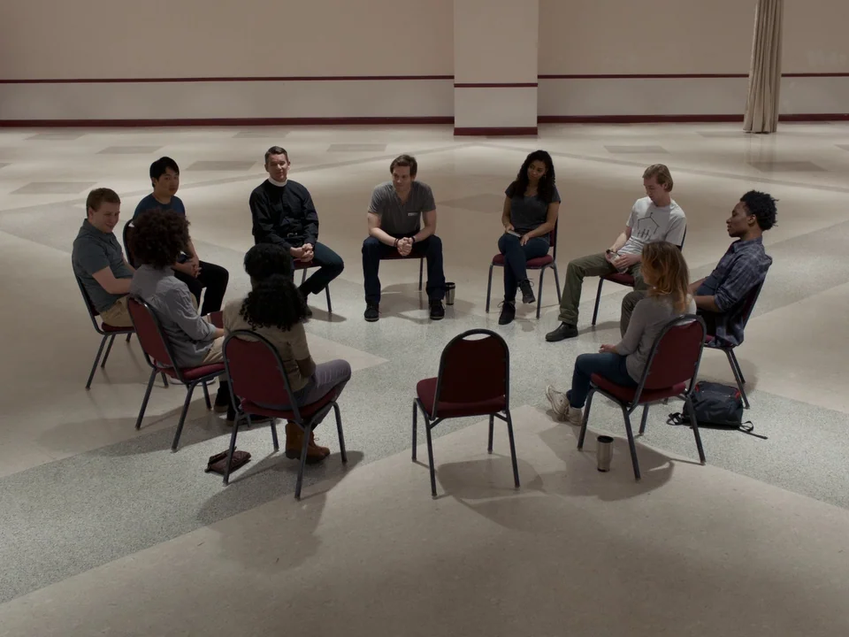 First Reformed (2017) movie still: wide — A wide, sparsely furnished community room features a circle of chairs on a tile floor. Nine diverse people…