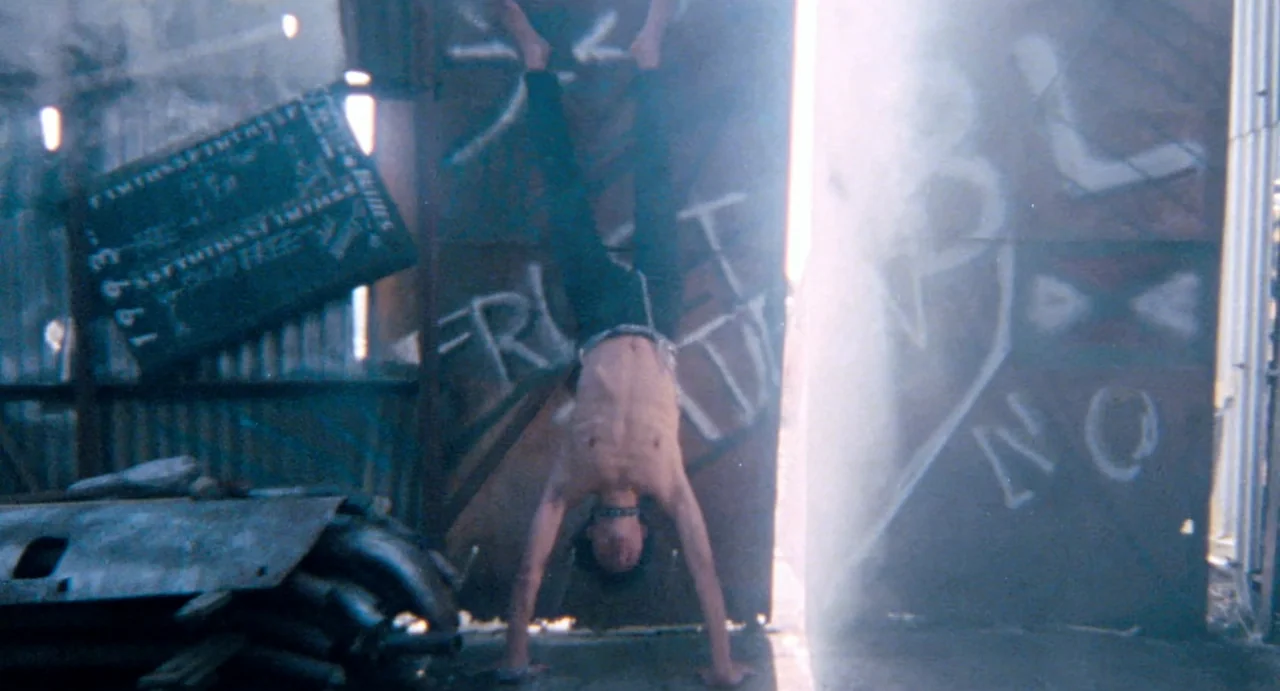 Burst City (1982) movie still: fullbody — An urban industrial area features metal walls covered in faded spray-painted graffiti tags. A shirtless…