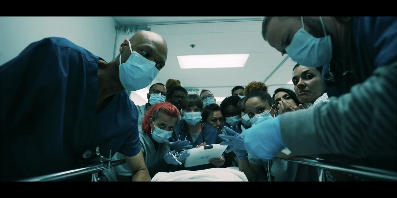Música (2024) movie still: medium — A hospital staff clusters around a patient perspective while looking down at a clipboard. The group wears blue…